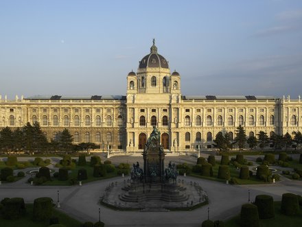 Das Bild zeigt die prächtige Fassade eines historischen Museums mit einer großen Kuppel. Im Vordergrund befindet sich ein schmuckvoller Brunnen und gepflegte Grünflächen mit Sträuchern. Der Himmel ist klar und die Sonne beleuchtet die Architektur.