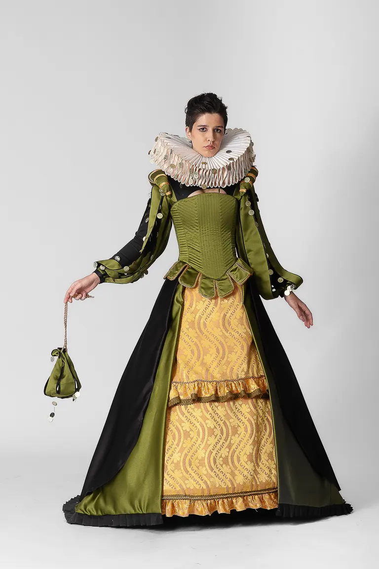 A model poses in a historical costume featuring a fitted green bodice, a wide pleated collar, and a long, flowing skirt with intricate yellow patterns. The ensemble is complemented by black accents and a small purse held in one hand, set against a plain white background.