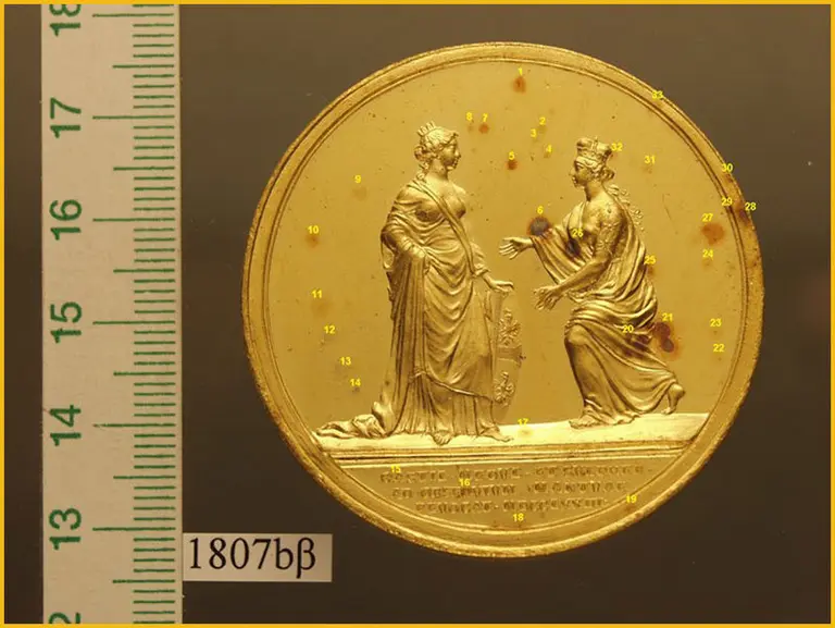 Die Abbildung zeigt eine goldene Medaille mit zwei weiblichen Figuren. Die linke Figur steht höflich, während die rechte Figur eine Hand ausstreckt. Im Hintergrund sind ornamental gestaltete Elemente zu sehen. Unterhalb der Figuren befindet sich eine Inschrift. Links ist ein Maßstab angedeutet.