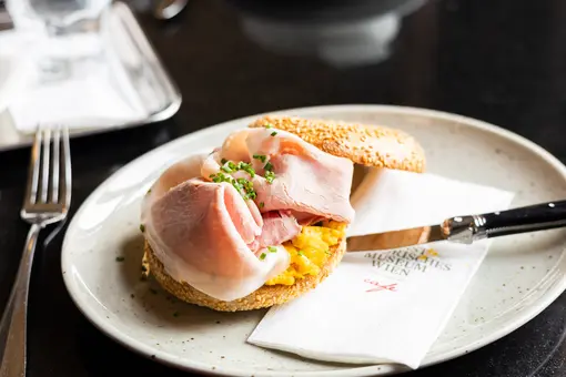 Ein Teller mit einem Brötchen, gefüllt mit Rührei und Scheiben von Schinken. Das Brötchen hat eine Sesamkruste und ist mit frischen Schnittlauch bestreut. Neben dem Teller liegt eine Serviette und ein Besteckset.
