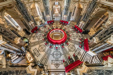 Eine Aufsicht auf einen prächtigen Innenraum mit einem runden, roten Tisch in der Mitte, umgeben von roten Stühlen. Säulen aus Marmor und ein kunstvolles Fußbodenmuster verleihen dem Raum Eleganz. Helle Lichtstrahlen fallen durch die oberen Öffnungen.