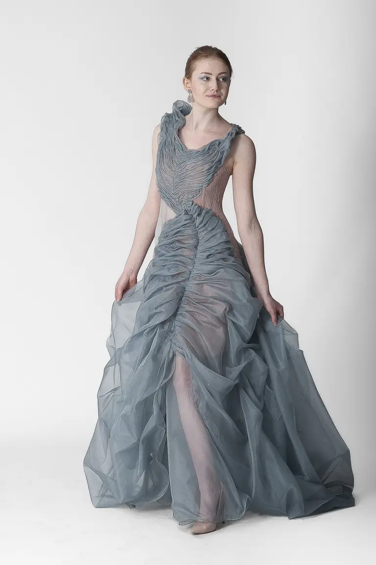 A model poses in an elegant, flowing gown made from sheer fabric. The dress is a soft blue-grey, featuring intricate draping and a ruffled neckline. The gown has a fitted bodice and a skirt that cascades gracefully to the floor. The background is a plain white.