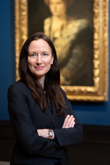 Eine Frau mit langen dunklen Haaren trägt einen schwarzen Blazer und steht mit verschränkten Armen vor einem Gemälde im Hintergrund. Der Raum ist in gedeckten Farben gehalten, und das Gemälde ist in einem goldenen Rahmen.