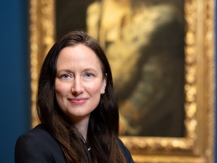 A woman with long dark hair stands confidently with her arms crossed, wearing a black blazer. She is positioned in front of a portrait featuring a historical figure, which is framed in gold. The background is a deep blue.