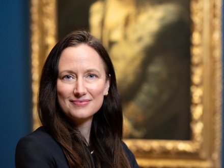 Eine Frau mit langen dunklen Haaren trägt einen schwarzen Blazer und steht mit verschränkten Armen vor einem Gemälde im Hintergrund. Der Raum ist in gedeckten Farben gehalten, und das Gemälde ist in einem goldenen Rahmen.