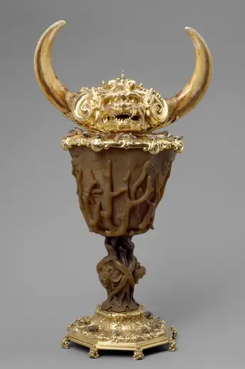 Ein kunstvoll gestalteter Becher aus goldfarbenem Material mit zwei großen, geschwungenen Hörnern und einem verzierten oberen Rand. Die äußere Form des Bechers zeigt eine Textur, die an Baumstämme erinnert, während die Basis mit einem filigranen, verzweigten Design verziert ist.
