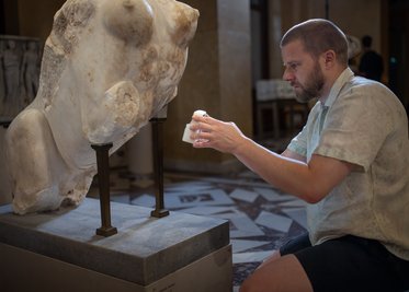 Ein Mann sitzt vor einer antiken Skulptur, die auf einem Standfuß platziert ist. Er hält ein kleines Objekt in seinen Händen und untersucht die Skulptur aufmerksam. Im Hintergrund sind unscharfe Konturen anderer Kunstwerke und Besucher zu sehen.