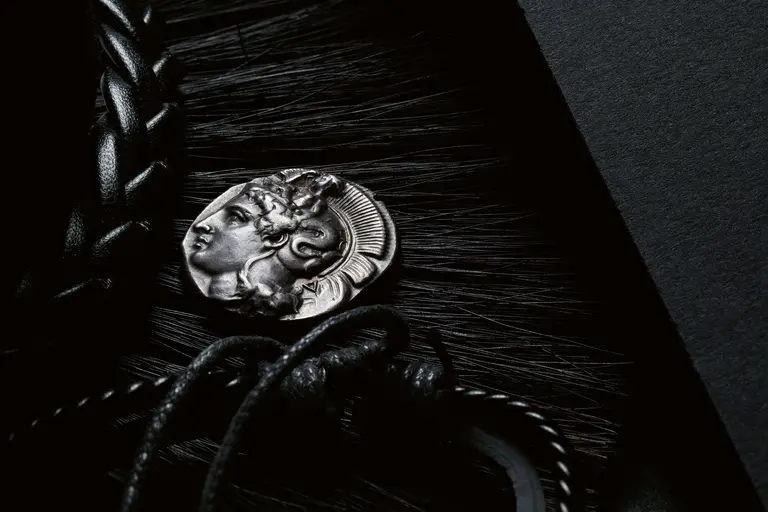 A close-up of a silver medallion featuring a detailed relief of a woman's face, set against a textured dark background. The medallion is partially draped with braided black leather, enhancing its antique appearance and elegance.