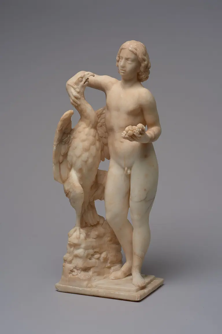 Eine Skulptur zeigt einen jungen Mann, der nackt vor einem großen Adler steht. Der Mann hält eine Hand an den Kopf des Adlers und trägt in der anderen Hand Früchte. Die Statue besteht aus marmorähnlichem Material und steht auf einem rechteckigen Sockel.
