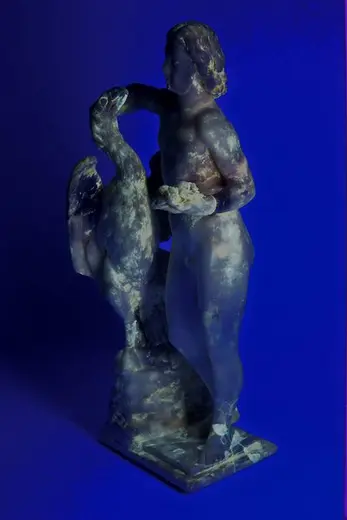 Die Skulptur zeigt eine menschliche Figur, die einen großen Vogel in der linken Hand hält und mit der rechten Hand dessen Kopf berührt. Die Figur ist detailliert gestaltet und steht auf einem Sockel, während der Hintergrund in einem kräftigen Blau gehalten ist.