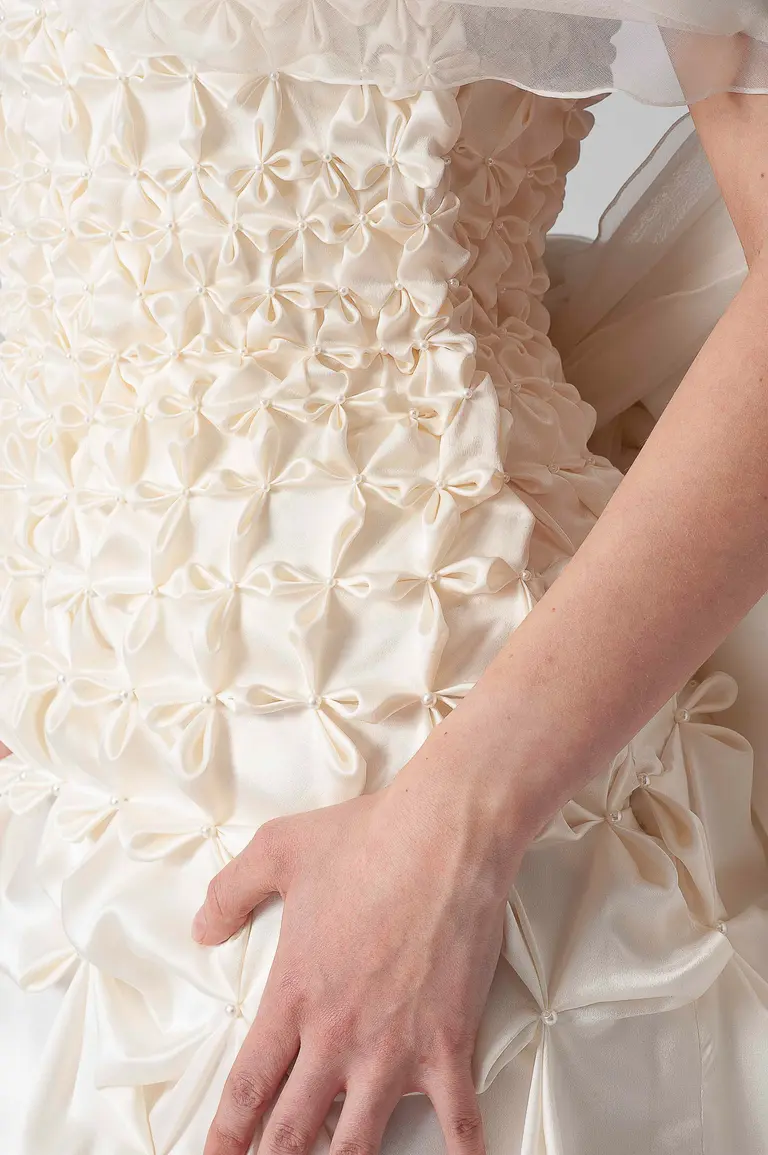 Close-up of a textured ivory wedding dress featuring intricate ruffles and bow-like embellishments. A hand rests on the dress, showcasing the delicate fabric and design details. The image focuses on the intricate craftsmanship of the dress.