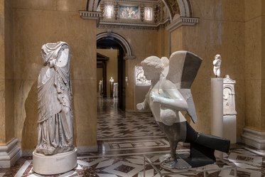 In einem Kunstmuseum sind zwei Skulpturen zu sehen: eine antike Statue einer weiblichen Figur links und eine moderne, abstrakte Figur eines geflügelten Wesens rechts. Der Raum ist mit marmorierten Fußböden und dekorativen Wänden gestaltet, während weitere Kunstwerke im Hintergrund angedeutet werden.