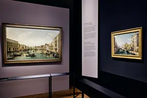 Zwei Landschaftsgemälde sind in einem Ausstellungsraum zu sehen. Links ein größeres Bild mit einer venezianischen Kanalansicht und Booten, rechts ein kleineres Gemälde mit einer ähnlichen Szene. An der Wand befindet sich eine Beschreibung zu den Kunstwerken.
