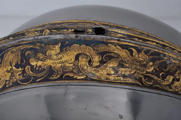 Eine detailreiche Metalloberfläche mit einem goldenen, spiralförmigen Muster, das floral und ornamental wirkt. Der Rand zeigt filigrane Verzierungen, die einen historischen oder kunsthistorischen Bezug vermuten lassen. Der Hintergrund ist schlicht, was den Fokus auf die dekorativen Elemente lenkt.