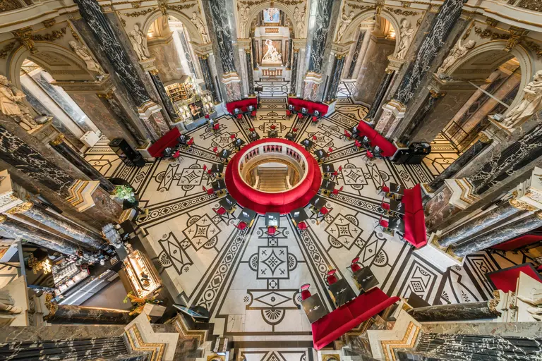 Der Blick von oben zeigt einen eleganten Innenraum mit einem kreisförmigen Tisch in der Mitte, umgeben von roten Stühlen. Die Bodenverzierung ist detailreich und geometrisch, umgeben von hohen Säulen und beeindruckenden architektonischen Elementen.