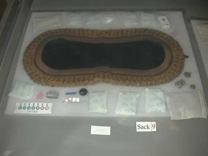 Die Abbildung zeigt ein ovales, mit einer goldenen Bordüre versehenes Tuch, das in einer Vitrine liegt. Um das Tuch herum sind mehrere kleine Tüten mit verschiedenen Inhalten angeordnet. Die Tüten sind auf einem weißen Untergrund platziert, auf dem auch Beschriftungen zu sehen sind.