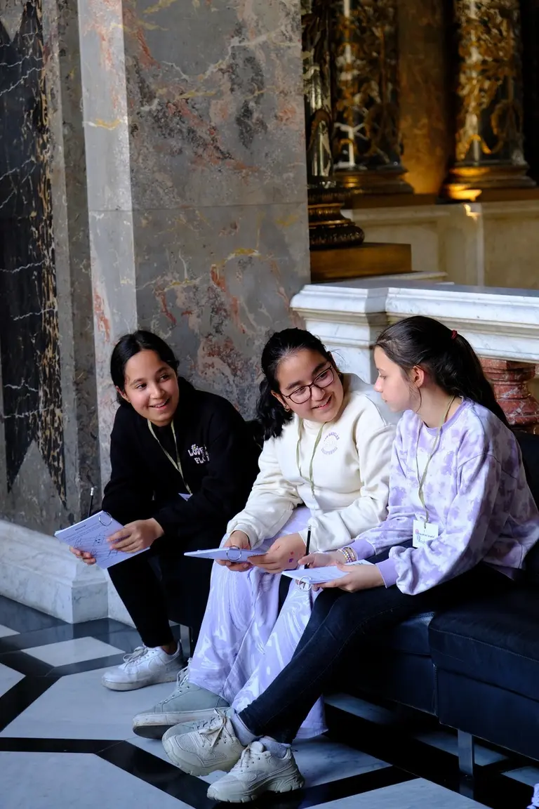 Drei Mädchen sitzen auf einer Bank in einem eleganten Raum. Sie unterhalten sich fröhlich und halten Blätter in der Hand. Die Umgebung ist reich verziert mit marmorartigen Wänden und symbolisiert eine kulturelle oder historische Stätte.