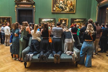 Mehrere Menschen stehen an einer Couch in einer Kunstgalerie und betrachten Gemälde an der Wand. Die meisten Personen sind gekleidet, als würden sie eine Ausstellung besuchen, und haben verschiedene Frisuren und Stile. Die Wände sind in einem grünen Farbton gestrichen.