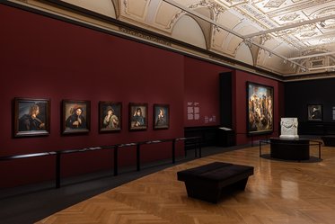 Ein Ausstellungsraum mit einer rot gestrichenen Wand, an der mehrere Bilder in schwarzen Rahmen hängen. Auf der gegenüberliegenden Seite befindet sich ein großer Gemälde sowie ein Tisch mit einer Skulptur in der Mitte des Raumes. Der Boden ist mit Holzpaneelen ausgelegt.