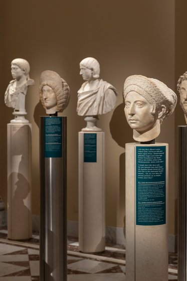 In einem Museum sind mehrere klassische Büsten auf weißen Sockeln ausgestellt. Jede Büste zeigt eine andere Person, dargestellt in verschiedenen Stilen. Informationsschilder an den Sockeln geben detaillierte Erläuterungen zu den Exponaten. Der Raum hat eine elegante, historische Atmosphäre.