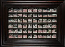 Das Bild zeigt eine große, dunkle Holzrahmenkonstruktion mit 81 kleinen, rechteckigen Bildern. Jedes Bild stellt verschiedene Szenen dar, die größtenteils in Rosa und Weiß gehalten sind, und illustrieren mythologische oder kunstvolle Darstellungen. Die Anordnung ist ordentlich und symmetrisch.