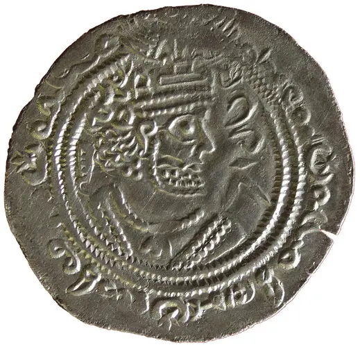 Der Bildinhalt zeigt eine silberne Münze mit dem Portrait eines gekrönten Herrschers, der einen Bart trägt. Um das Porträt herum sind kunstvolle Verzierungen und ein Randmuster zu sehen. Die Münze vermittelt einen historischen Kontext und könnte aus einer älteren Zivilisation stammen.