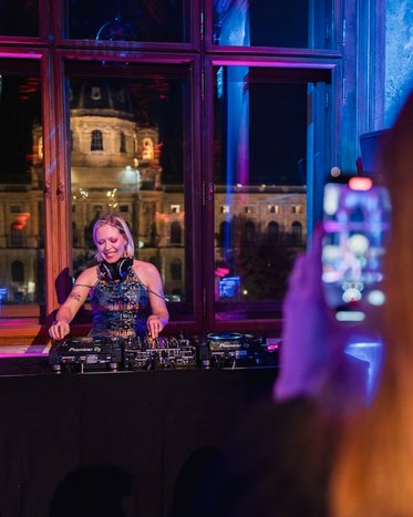 Eine DJane steht hinter einem DJ-Pult und mischt Musik. Im Hintergrund sind große Fenster mit einem Blick auf ein beleuchtetes Gebäude. Eine Person hält ein Handy und nimmt die DJane auf. Die Atmosphäre ist festlich und bunt beleuchtet.