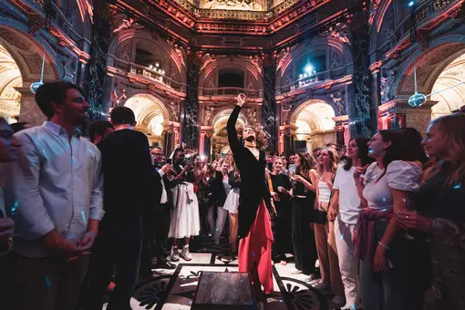 A crowd of people watches an expressive drag performer at an event in the Cupola Hall. The performer poses while the audience, holding drinks and capturing moments on their phones, enjoys the vibrant atmosphere.