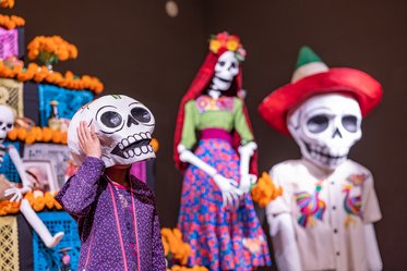 Ein Kind trägt eine Skeletthutmaske und steht vor einer farbenfrohen, traditionellen mexikanischen Dekoration mit Blumen und Figuren von Skeletten in festlicher Kleidung. Die Szenerie feiert das Día de los Muertos, mit lebhaften Farben und festlicher Atmosphäre.