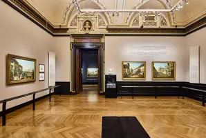 Ein Kunstmuseum mit hohen Decken und einem eleganten Boden. An den Wänden hängen verschiedene Gemälde, darunter Landschaftsdarstellungen. Ein Türöffnung führt zu weiteren Exponaten im Hintergrund. Informationen sind in Textform an der Wand angebracht.