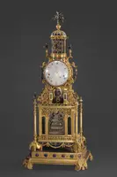 Elegante goldene Tischuhr mit einem runden Ziffernblatt, verziert mit filigranen Details und bunten Edelsteinen. Oben befindet sich ein kunstvolles Gitterwerk, das einen dekorativen Abschluss bildet. Die Uhr erinnert an einen historischen Stil und ist ein Beispiel für kunsthandwerkliche Meisterschaft.