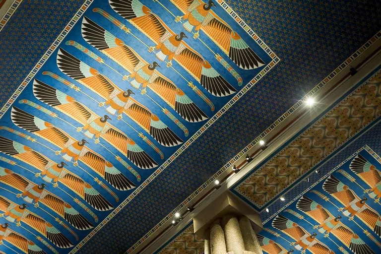Decke mit kunstvollen blauen und goldenen Mustern, die geflügelte Sonnenzeichen darstellen. Die Farben sind lebendig und der Raum wirkt festlich. Die Wand- und Deckengestaltung zeigt ägyptische Einflüsse und ist reich an Details.