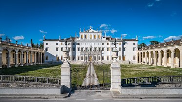 Eine Ansicht eines eleganten, historisch architektonischen Gebäudes mit weißen Wänden und einem gepflegten Garten. Das Gebäude hat eine zentrale Fassade mit Säulen und Verzierungen, flankiert von niedrigen Gebäuden in einem Innenhof. Der Himmel ist klar und blau.