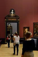 Besucher erkunden eine Kunstgalerie mit roten Wänden. Im Hintergrund ist ein Spiegel mit einer Büste darüber zu sehen, während Gemälde an den Wänden hängen. Die Menschen gehen in Richtung des Spiegels, was eine Atmosphäre von Neugier und Erkundung vermittelt.