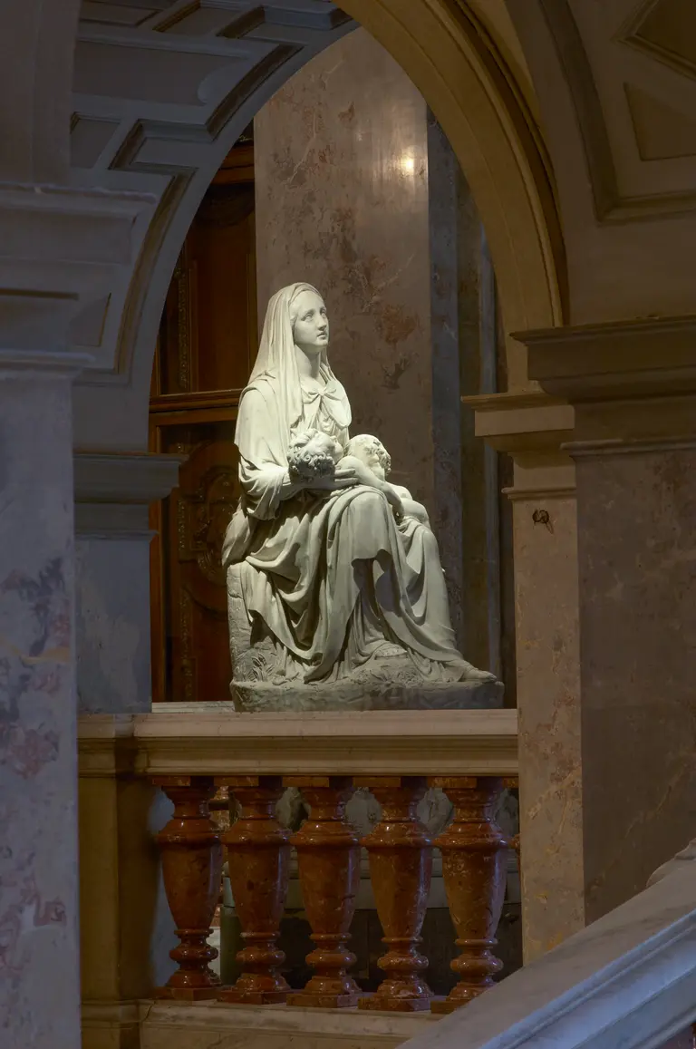Die Skulptur zeigt eine sitzende Figur einer Frau, die ein Kind auf dem Schoß hält. Der Ausdruck der Frau vermittelt Ruhe und Fürsorge. Die Statue ist in einem hellen Material gefertigt und steht in einem architektonisch gestalteten Raum, umgeben von Säulen und Wänden.
