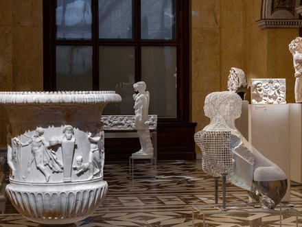 Eine Kunstinstallation im Museum zeigt eine weiße Marmorschale mit Reliefs von Figuren, eine skulpturierte menschliche Figur und eine durchsichtige, abstrahierte Figur. Die Werke sind auf Sockeln platziert und vor einem fensterähnlichen Hintergrund aus Holz.