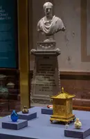 Im Vordergrund sind drei dekorierte Fläschchen in verschiedenen Farben und eine goldene, quadratische Kiste auf einem Tisch zu sehen. Im Hintergrund steht eine Büste einer antiken Figur, die in einem Museum ausgestellt ist.