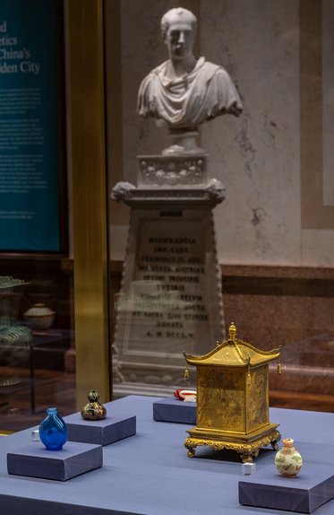 Im Vordergrund sind drei dekorierte Fläschchen in verschiedenen Farben und eine goldene, quadratische Kiste auf einem Tisch zu sehen. Im Hintergrund steht eine Büste einer antiken Figur, die in einem Museum ausgestellt ist.