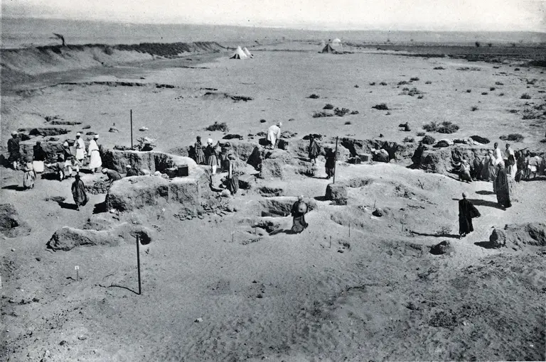 Historisches Schwarz-Weiß-Foto der archäologischen Ausgrabung aus dem Jahr 1910 in einer wüstenartigen Landschaft. Menschen arbeiten an der Freilegung von Erd- und Steinformationen, die vermutlich Teil der antiken Siedlung sind. Im Hintergrund kleine Zelte und weite, flache Ebene.