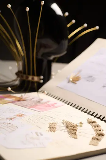 A black mannequin head adorned with decorative golden pins sits beside an open sketchbook. The pages contain various designs, notes, and dried flowers, showcasing creative ideas and fashion concepts. The overall focus highlights artistry in fashion design and inspiration.