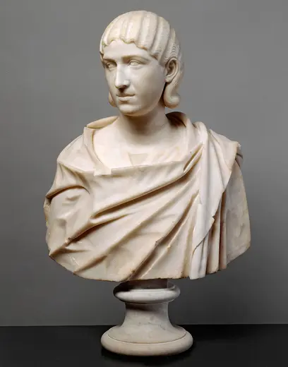 Eine antike Marmorbüste eines jungen Mannes mit lockigem Haar. Er trägt eine drapierte Tunika und blickt sanft nach vorne. Die Büste ruht auf einem runden Sockel und vermittelt einen Eindruck von klassischer Schönheit und Eleganz.