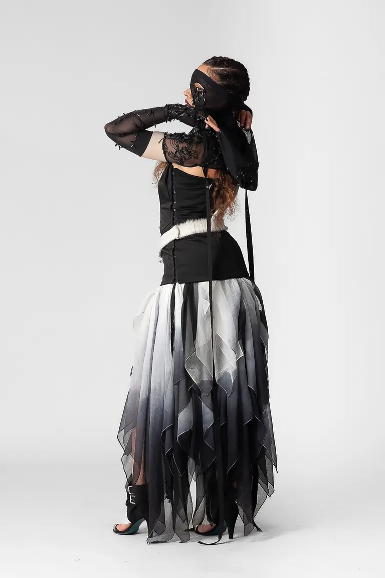 A model stands with her back facing the camera, wearing a unique outfit that features a layered, ombre skirt transitioning from white to black. The top is form-fitting with sheer black sleeves. She has her hands raised near her face, partially obscured by a veil.