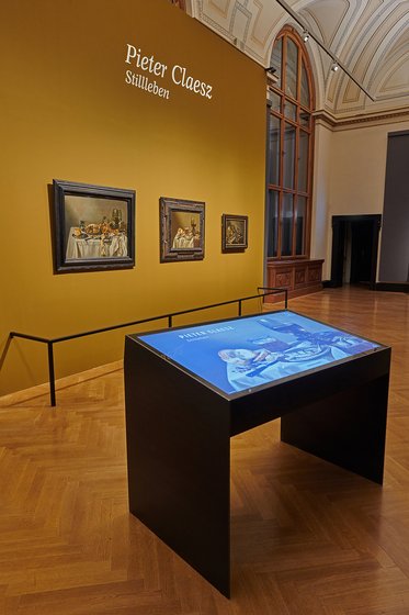 Das Bild zeigt eine Kunstausstellung mit dem Titel "Pieter Claesz - Stillleben". An der Wand sind mehrere Stillleben-Gemälde zu sehen, während ein Tisch mit interaktiver Präsentation im Vordergrund steht. Der Raum hat hohe Fenster und einen eleganten Holzfußboden.