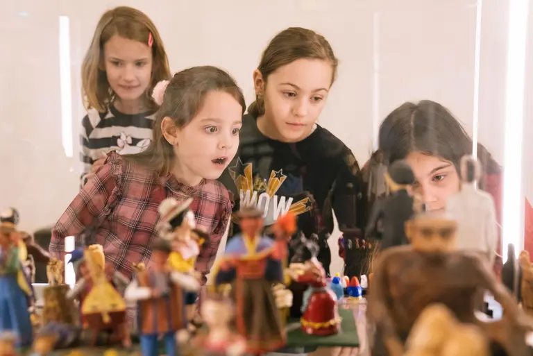 Vier Mädchen schauen neugierig auf eine Ausstellung von bunten Figuren in einem Glaskasten. Ihre Gesichter zeigen Staunen und Begeisterung für die dargestellten Objekte.