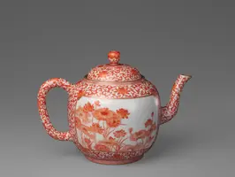 Eine Teekanne aus Porzellan mit einer dekorativen roten Musterung von Blumen und einem geschwungenen Auslauf. Der Deckel hat einen runden Knauf. Die Kanne hat einen breiten Griff und ist auf einem grauen Hintergrund platziert.