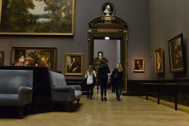 Eine Gruppe von Personen geht in einem großen Ausstellungsraum eines Kunstmuseums. An den Wänden hängen verschiedene Gemälde, und im Hintergrund ist eine große Tür zu sehen, die den Zugang zu einem weiteren Raum ermöglicht. Der Boden ist aus Holz.
