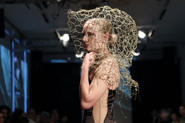 Eine Frau läuft auf einem Laufsteg und trägt ein elegantes, dunkles Kleid. Ihr Kopf ist mit einem kunstvollen, netzartigen Kopfschmuck geschmückt. Der Hintergrund zeigt eine aufmerksame Menge in gedämpftem Licht.