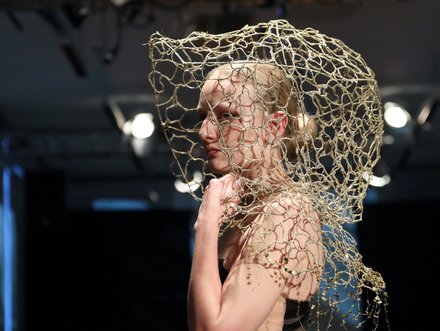 Eine Frau läuft auf einem Laufsteg und trägt ein elegantes, dunkles Kleid. Ihr Kopf ist mit einem kunstvollen, netzartigen Kopfschmuck geschmückt. Der Hintergrund zeigt eine aufmerksame Menge in gedämpftem Licht.