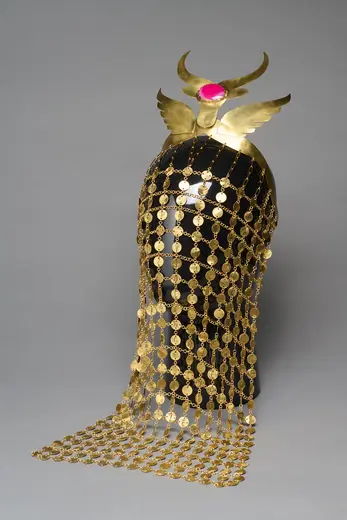 A decorative headpiece made of gold and adorned with a pink gem, featuring intricate chains of gold coins cascading down. The design includes ornate wings at the top, suggesting a blend of elegance and cultural significance. The background is neutral, highlighting the craftsmanship of the piece.