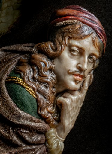 Eine detailreiche Skulptur eines Mannes mit langen, gewellten Haaren und einem roten Kopftuch. Er hat einen nachdenklichen Gesichtsausdruck, während er mit einem Finger nachdenklich sein Kinn berührt. Der Mann trägt ein grünes Gewand mit goldenen Verzierungen.